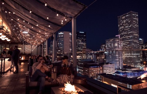A rooftop bar at night with string lights, people socializing, and a city skyline in the background.