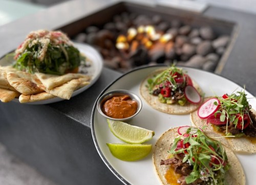 A plate of tacos with toppings, lime wedges, and a side of salad and flatbread, set near a fire pit.