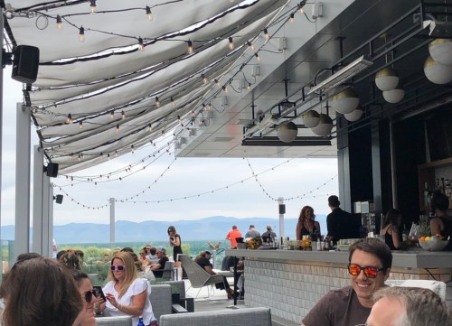 A rooftop bar with string lights, people socializing, and a scenic view of mountains in the background.