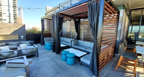 Rooftop lounge area with modern seating, blue stools, and curtains, surrounded by city views and buildings.