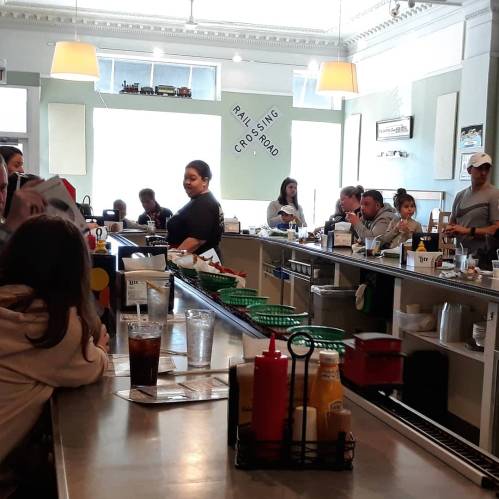 A busy diner scene with customers at the counter, a waitress serving, and drinks on the bar. Bright and inviting atmosphere.