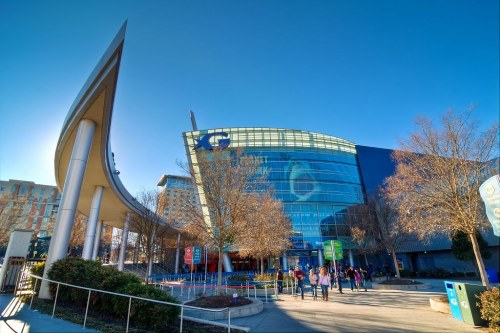 A modern building with a curved roof and large glass windows, surrounded by trees and people walking outside.