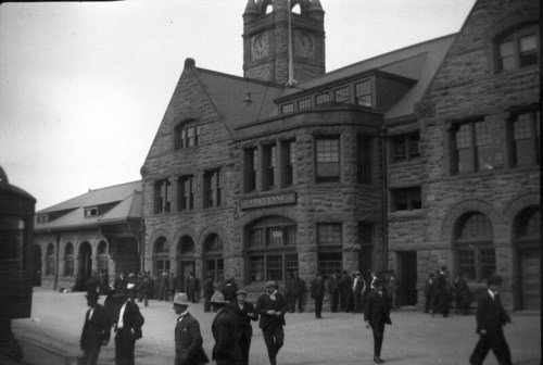 Historic train station with stone architecture, clock tower, and crowds of people in early 20th-century attire.