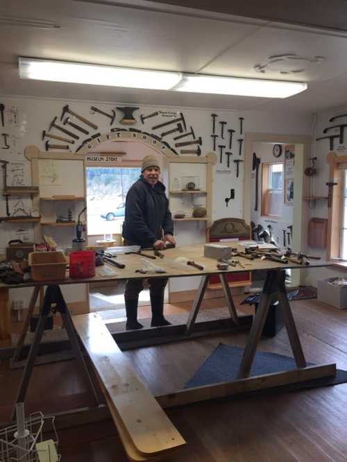 A person stands at a workbench in a workshop filled with tools, smiling and surrounded by various woodworking equipment.