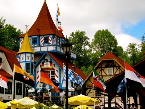 Colorful buildings with a tall, pointed tower, surrounded by flags and outdoor seating under cloudy skies.