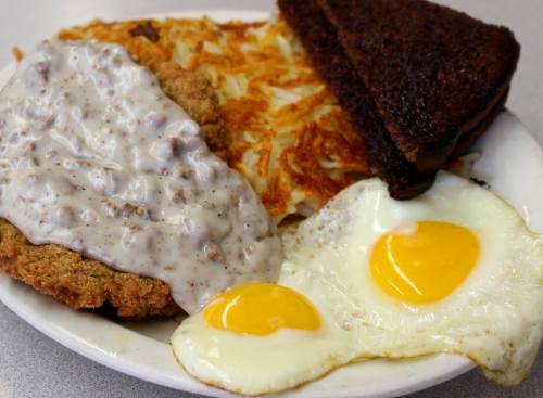 A plate of breakfast featuring fried chicken with gravy, hash browns, two sunny-side-up eggs, and toasted bread.