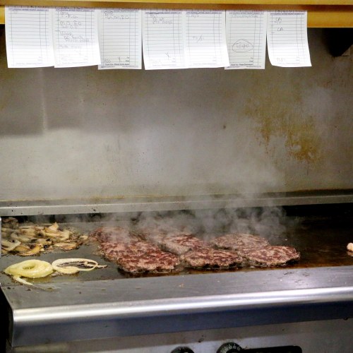 Grill with sizzling burgers and onions, steam rising, and order tickets hanging above.