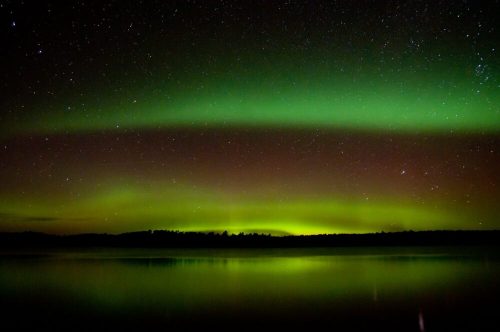 A serene night sky illuminated by green auroras reflecting on a calm lake, surrounded by dark silhouettes of trees.