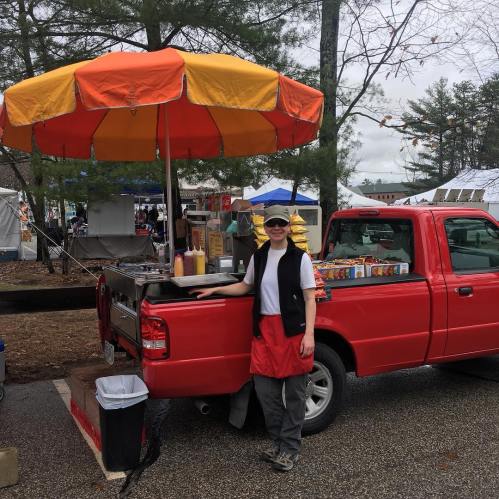 A vendor stands by a red food truck with an orange umbrella, serving food at an outdoor market.