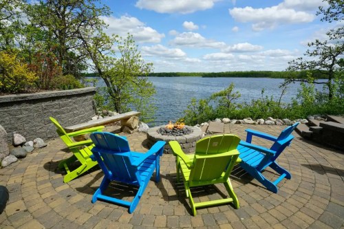 Colorful adirondack chairs surround a stone fire pit by a serene lake, with trees and clouds in the background.