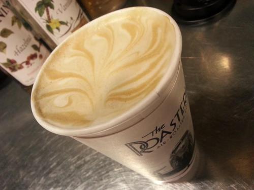 A close-up of a latte in a paper cup, featuring intricate foam art on top.