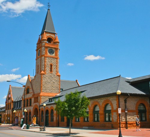 Historic stone building with a tall clock tower, set against a clear blue sky. Trees and street lamps line the sidewalk.