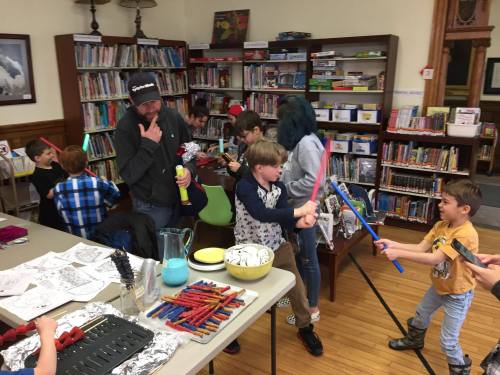 A lively library scene with children playing, crafting, and engaging in activities, while an adult observes thoughtfully.