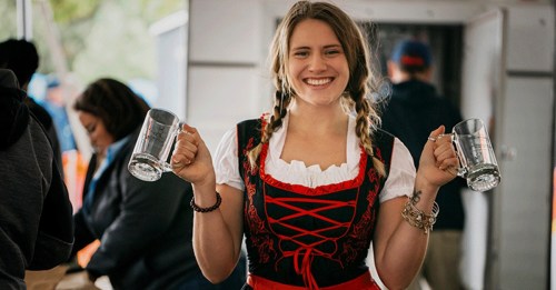 A smiling woman in a traditional dress holds two beer mugs, standing in a festive outdoor setting.