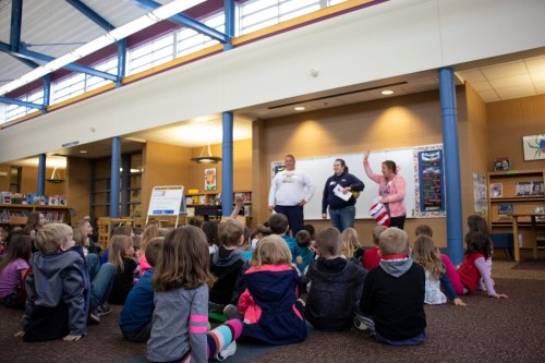 A group of children sits on the floor in a library, listening to three adults speaking at the front.