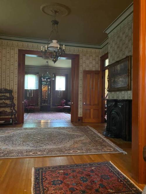 A vintage room with patterned wallpaper, a chandelier, and ornate furnishings, featuring a large area rug on wooden floors.