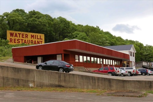 A red and white building with large windows, labeled "Water Mill Restaurant," surrounded by parked cars and greenery.