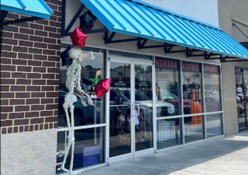 A storefront with blue awnings, featuring a skeleton holding balloons and a display of horror-themed apparel inside.