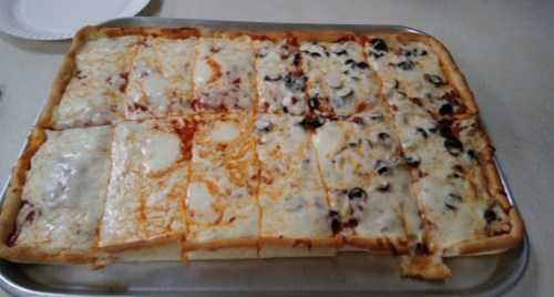 A rectangular tray of pizza with various toppings, including cheese, sauce, and olives, cut into square slices.