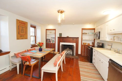 Cozy kitchen with a dining table, colorful chairs, a fireplace, and white cabinets, featuring warm wooden floors.
