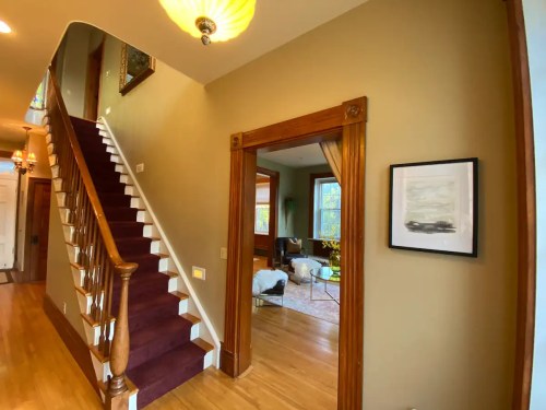 A hallway with a staircase, wooden trim, and a doorway leading to a cozy living room with natural light.