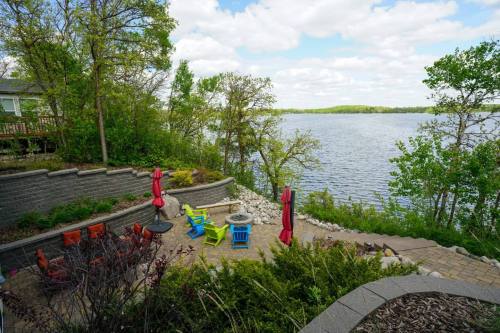 A scenic view of a lakeside patio with colorful chairs, umbrellas, and lush greenery under a partly cloudy sky.