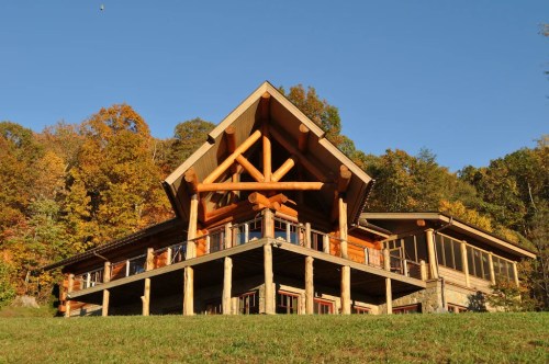 A large log cabin with a peaked roof, surrounded by colorful autumn trees and a clear blue sky.