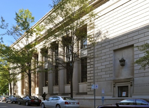A large, classical-style building with tall columns, surrounded by trees and parked cars on a sunny day.