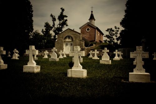 A cemetery with white crosses marking graves, set against a backdrop of a church and trees under a cloudy sky.