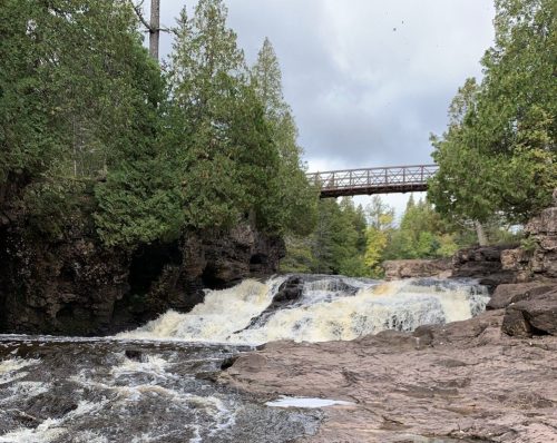 A scenic view of a waterfall surrounded by trees, with a bridge in the background and rocky terrain in the foreground.