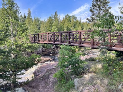 A wooden bridge spans a rocky stream, surrounded by lush green trees and a clear blue sky.