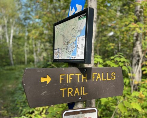 Trail sign for Fifth Falls Trail with a map, surrounded by greenery.