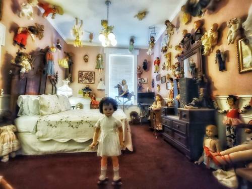 A vintage bedroom filled with dolls on the walls and furniture, featuring a bed and a central doll in a white dress.