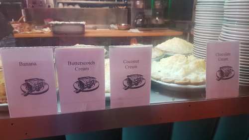 Four dessert signs display: Banana Cream, Butterscotch Cream, Coconut Cream, and Chocolate Cream pies in a bakery.