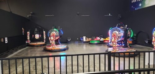 A game arcade featuring colorful spinning rides with glowing lights, set in a spacious, dark room.