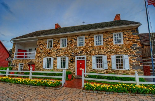 A historic stone house with red doors and white trim, surrounded by colorful flowers and a picket fence.