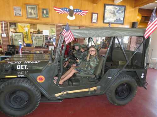 A military jeep with flags, occupied by children in camouflage, inside a decorated room with patriotic memorabilia.