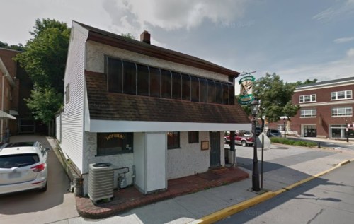 A small, two-story building with a sloped roof and large windows, located on a street corner with parked cars nearby.