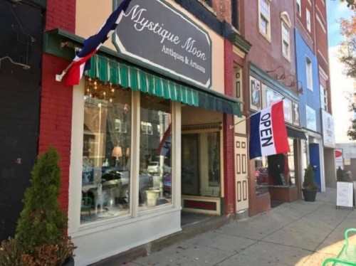 A quaint antique shop with a green awning and "OPEN" sign, flanked by colorful buildings and flags.