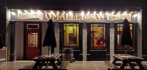 A storefront with the sign "O'Malley & Son Wine & Spirits," featuring outdoor seating and illuminated signage.