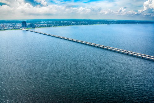 Aerial view of a long bridge extending over a calm body of water, with a city skyline in the background under a blue sky.