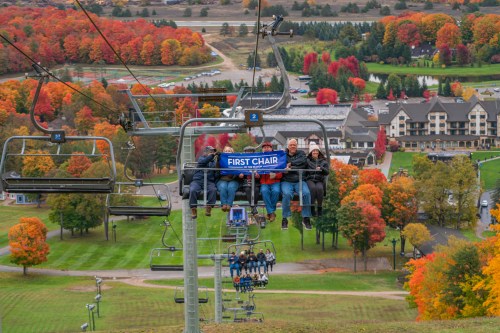 A group of four people on a ski lift, surrounded by vibrant autumn foliage and a scenic resort below.