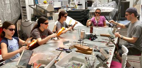 A group of five people wearing sunglasses work on glassblowing projects at a table filled with tools and materials.