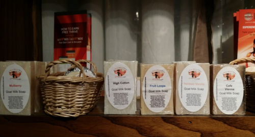 A display of goat milk soaps in various scents, with a woven basket on a wooden shelf.