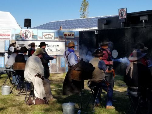A group of people in Western attire participate in a shooting competition outdoors, with smoke rising from their guns.