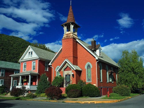 A red brick church with a steeple, surrounded by greenery and a nearby house, under a blue sky with clouds.