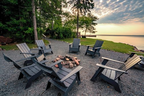 A circle of black Adirondack chairs around a fire pit by a serene lakeside at sunset.