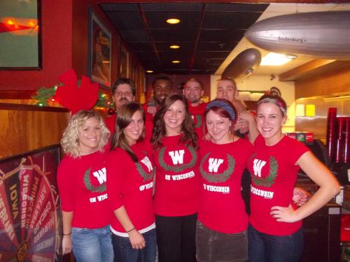 A group of people in festive red shirts posing together in a decorated restaurant, smiling and celebrating.