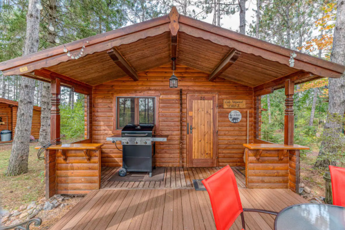 Cozy wooden cabin with a grill on the porch, surrounded by trees and a wooden deck with red chairs.