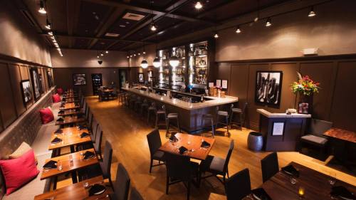 A modern restaurant interior featuring wooden tables, a bar, and stylish decor with dim lighting and framed photos.